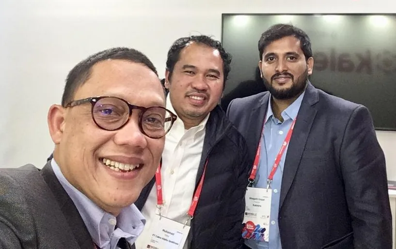 Tiga pria tersenyum berfoto selfie di depan stan pameran MWC Barcelona, mengenakan tanda pengenal acara