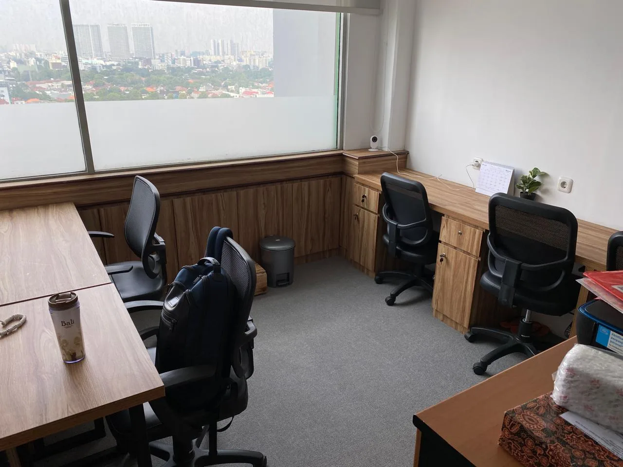 Ruang kantor kecil dengan meja kayu di sepanjang dinding, kursi kantor hitam, dan jendela yang menghadap gedung-gedung Jakarta